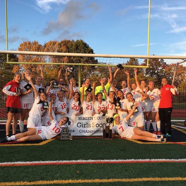 A girls' soccer team mid-cheer at golden hour, arms up around the Bergen County championship trophy