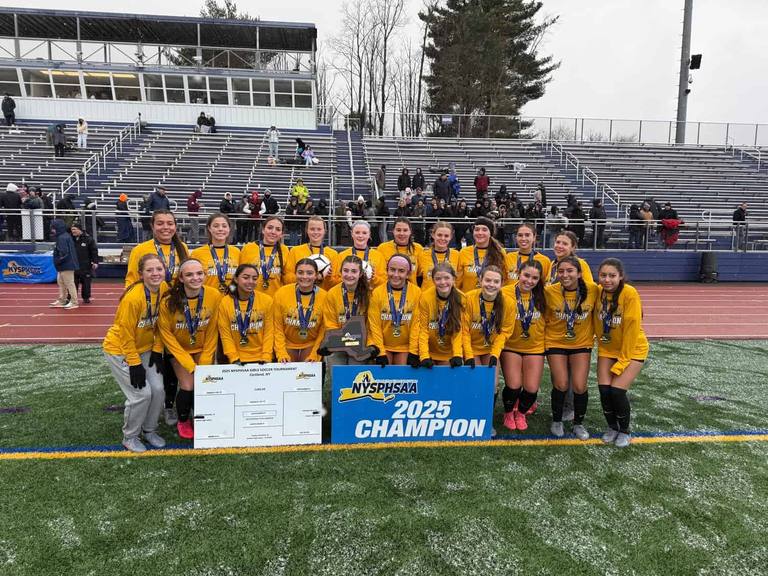 A girls’ soccer team on the field with their 2025 state champion trophy and banner