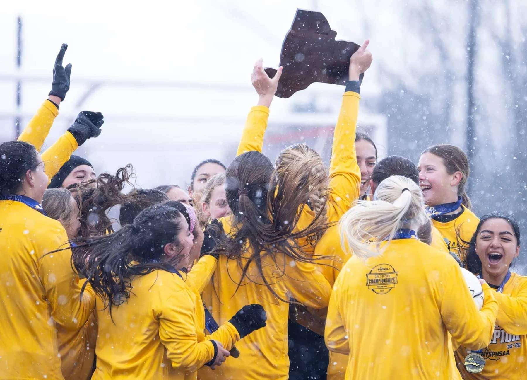 Girls' soccer team hoisting a state-championship trophy in falling snow
